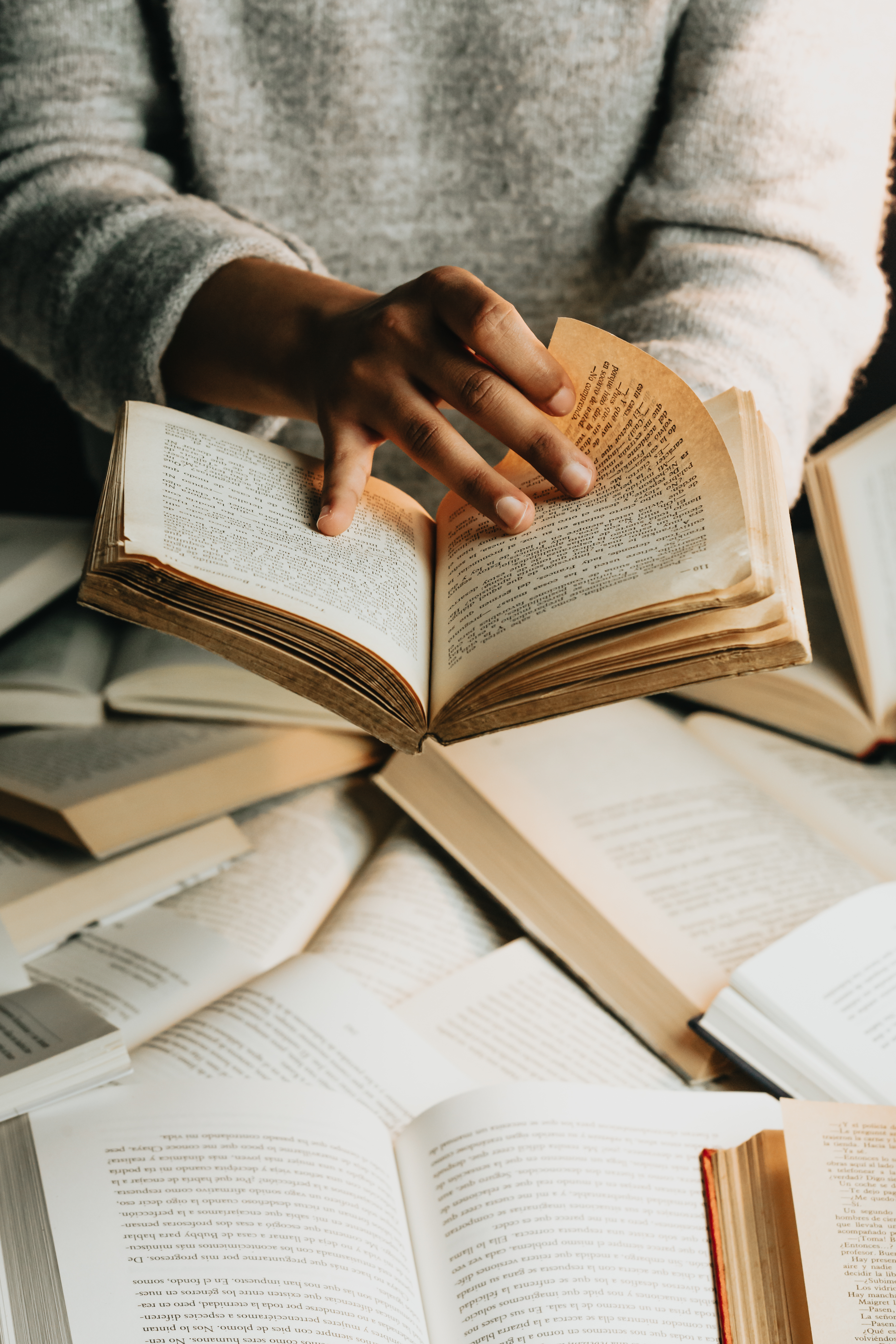Hands reading a book with a pile of open books underneath